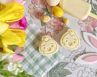 Op de afbeelding: Twee witte chocolade kip koekjesvormen op een groen-wit geblokt doek met een bloemige achtergrond. Er zijn ook verschillende gele en witte paaseieren en een witte chocolade reep in de afbeelding.