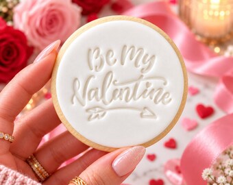 May include: A round sugar cookie with smooth white icing featuring the embossed text Be My Valentine and a small arrow graphic. The cookie is held against a background of red and pink roses, pink ribbon, and small red heart confetti.
