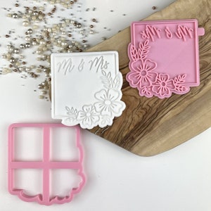 Peut inclure: Trois emportes-pièces en plastique rose et blanc. Un emporte-pièce est un carré avec les mots "Mr & Mrs" et un motif floral. Un autre emporte-pièce est un carré avec un motif floral. Le troisième emporte-pièce est un carré avec un motif de grille.