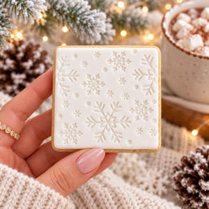 May include: A square cookie with a smooth white icing surface featuring an embossed pattern of various snowflake shapes. The cookie has a light golden brown edge and a flat, uniform design across the entire top surface.