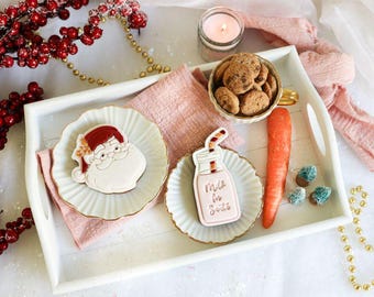 Puede incluir: Una bandeja blanca con detalles en rosa y dorado presenta una selección de galletas con temática navideña. Las galletas están decoradas con glaseado rojo y blanco y presentan diseños de Papá Noel y una botella de leche con el texto "Milk for Santa".