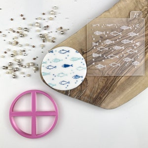 Pode incluir: Um biscoito redondo de fondant branco com um padrão de peixes azuis e verdes. Um estêncil de acrílico transparente com um padrão de peixes está sobre uma tábua de cortar de madeira. Um cortador de círculo de plástico rosa está sobre uma superfície branca.