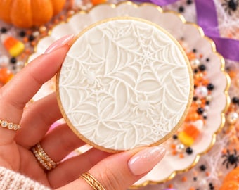 May include: A round sugar cookie topped with smooth white icing featuring an embossed spider web pattern and small spider figures. The cookie has a light golden brown edge and is held against a background of scattered Halloween candy.