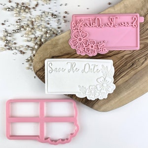 May include: Two pink and white plastic cookie cutters with the text "Save the Date" on them. The cutters are on a wooden surface.