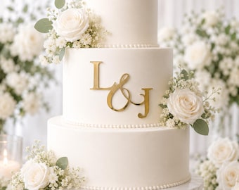 May include: A three-tier white cake with smooth frosting and a beaded border at the base of each tier. The middle tier features a gold-colored acrylic topper with the letters L and J connected by an ampersand. White roses and baby's breath decorate the tiers.