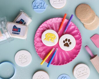 Puede incluir: Un plato rosa con una variedad de cortadores de galletas con forma de perro y galletas de fondant. El plato está rodeado de otros cortadores de galletas, fondant y un rodillo. El texto "Little Bake Box Fondant" es visible en un paquete blanco.