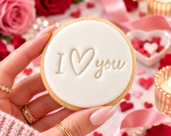 Puede incluir: Una galleta redonda cubierta con glaseado blanco liso que presenta el texto en relieve I heart you en color tostado claro. La galleta tiene un borde dorado y se muestra sobre un fondo de cintas rosas y pétalos de rosa rojos.