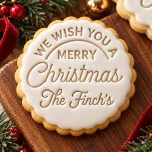 Puede incluir: Una galleta redonda de borde festoneado cubierta con glaseado blanco liso. El glaseado está grabado con el texto marrón "WE WISH YOU A MERRY Christmas The Finch's" en una mezcla de letras de bloque y cursivas, centrado en la superficie.