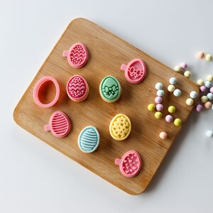 May include: A wooden board with nine Easter-themed cookie cutters. The cutters are in the shapes of eggs with different designs, including hearts, stripes, and polka dots. The cutters are in various colours, including pink, green, yellow, and blue.