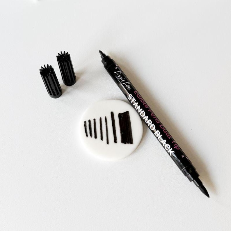 Puede incluir: Un rotulador comestible negro de doble punta con el texto Lissie Lou Edible Pens Dual Tip STANDARD BLACK impreso en el cuerpo, acompa&ntilde;ado de dos tapas negras y un disco circular blanco con una serie de l&iacute;neas verticales negras.