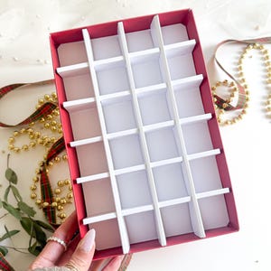 May include: A red box with white dividers, designed for compartmentalised storage. The box is divided into numerous small sections by a grid of white partitions. A tartan ribbon and gold beads are in the background, suggesting a festive theme.