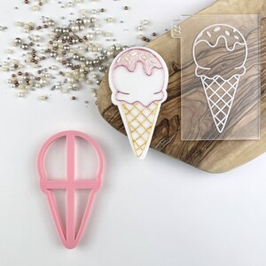 May include: Pink plastic ice cream cone cookie cutter with a waffle cone design. A white ice cream cone with pink and white frosting is shown next to the cutter. A clear plastic stencil of an ice cream cone is also shown.