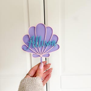 Puede incluir: Topper de pastel en forma de concha de color morado y rosa con el nombre Allison escrito en azul.