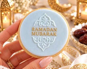 May include: A round sugar cookie with a smooth light blue icing surface. The center features an embossed white filigree design surrounding the gold-colored text "RAMADAN MUBARAK". The cookie has a golden-brown baked edge and a circular shape.
