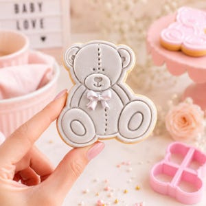 May include: A light gray teddy bear shaped cookie with a white icing bow at the neck, featuring stitched detailing and a subtle shimmer finish. The cookie is decorated with a heart shaped nose and rounded ears, presented as a single baked treat.
