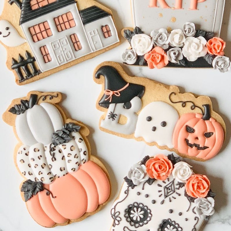 Puede incluir: Una colecci&oacute;n de galletas de az&uacute;car con tem&aacute;tica de Halloween que incluye una casa encantada con un fantasma, una l&aacute;pida con flores, una pila de calabazas con estampado de leopardo, la palabra BOO con un sombrero de bruja y una calavera.