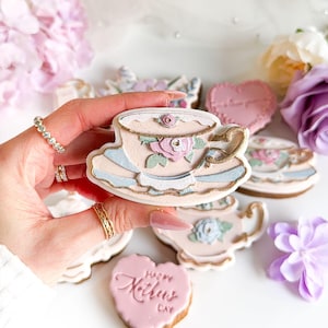 Könnte beinhalten: Eine Sammlung dekorierter Kekse in Form von Teetassen, Teekannen und Herzen. Die Kekse sind hauptsächlich pastellrosa, blau und goldfarben, mit floralen Designs. Ein herzförmiger Keks trägt die Aufschrift "Happy Mother's Day".