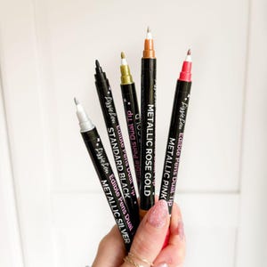 Puede incluir: Un juego de cinco rotuladores comestibles de doble punta LissieLou en plata met&aacute;lica, negro est&aacute;ndar, oro, oro rosa met&aacute;lico y rosa met&aacute;lico. Cada rotulador negro presenta texto en blanco que identifica el color y la marca.