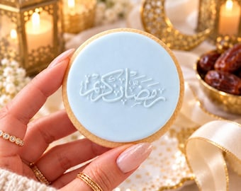 May include: A round sugar cookie with a smooth light blue icing layer featuring embossed white Arabic calligraphy that reads Ramadan Kareem. The cookie has a golden brown edge and is held against a background of gold lanterns and dates.