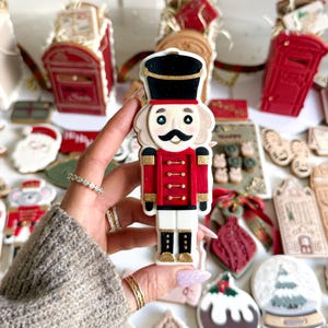 May include: A hand holds a decorated cookie shaped like a nutcracker soldier, with a black hat and red jacket. Other Christmas-themed cookies are visible, including a Santa, a snow globe, and a red postbox, all on a white surface.