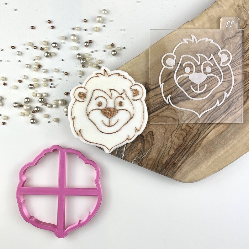 May include: A white fondant lion cookie with a gold glitter outline. The cookie is next to a clear plastic stencil of a lion face and a pink plastic cookie cutter with a cross-hatch design.