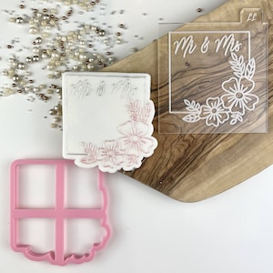 Peut inclure: Un pochoir en acrylique transparent avec le texte "Mr & Mrs" et un motif floral. Le pochoir est posé sur une surface en bois. Il y a également un moule en silicone rose avec un motif carré et une bordure florale.