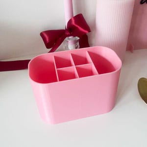 Puede incluir: Un organizador de escritorio de color rosa claro con forma ovalada y cuatro compartimentos internos de distintos tama&ntilde;os. El recipiente tiene paredes lisas y s&oacute;lidas con una parte superior abierta, ofreciendo un espacio estructurado.