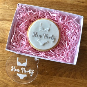 Puede incluir: Una galleta blanca con un diseño de encaje y el texto "Hen Party" hecho con glaseado plateado. La galleta está en una caja blanca con papel de seda rosa. También se incluye una plantilla de acrílico transparente con el mismo diseño.