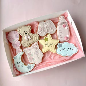 May include: A collection of decorated sugar cookies in a white box, featuring a beige teddy bear, pink baby booties, a baby bottle, a rattle, a onesie with floral details, a yellow star, a blue cloud, and a crescent moon with silver stars.