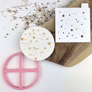 Pode incluir: Um conjunto de três ferramentas para decorar bolos: um estêncil branco com um padrão de estrelas, um cortador de círculo de silicone rosa com um design de cruz e um bolo redondo branco decorado com estrelas douradas.