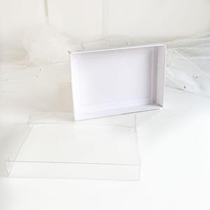 May include: A white cardboard box with a clear plastic lid. The box is empty and the lid is open.