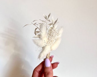 May include: A small boutonniere composed of dried white botanical elements, including fluffy bunny tail grass, delicate baby's breath, and slender leaves. The stems are tightly bound together with natural tan twine at the base of the arrangement.
