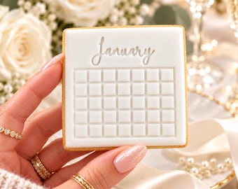 May include: A square cookie with white icing featuring the word January in cursive script above a grid pattern representing a calendar. The cookie has a light brown edge and is held against a background of white roses and ribbons.
