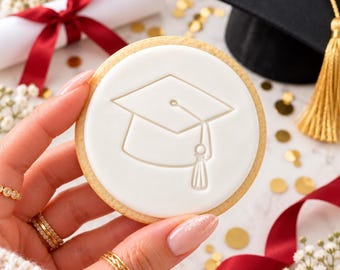 May include: A round sugar cookie topped with smooth white icing featuring an embossed graduation cap design. The cookie has a light golden brown edge and is held in place, showcasing the detailed outline of the mortarboard and tassel.