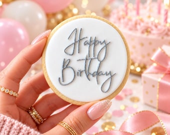 May include: A round sugar cookie with smooth white icing and the text Happy Birthday written in elegant grey script. The cookie is held against a background of pink and white balloons, a frosted cake with lit candles, and a gift box.