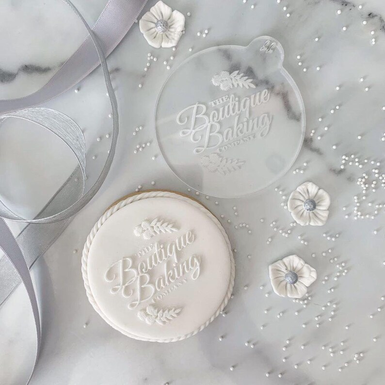 May include: White cookie with a white icing design that says "The Boutique Baking Company" with a floral design. A clear plastic cookie cutter with the same design is also shown.