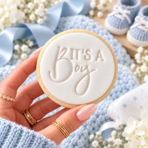 May include: A round sugar cookie topped with smooth white icing featuring the embossed text IT'S A BOY in a mix of block and script lettering. The cookie is held against a backdrop of light blue knitted fabric and baby booties.