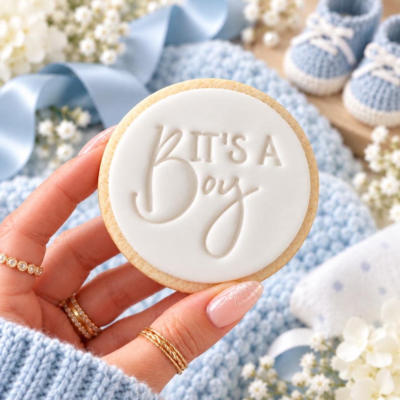 May include: A round sugar cookie topped with smooth white icing featuring the embossed text IT'S A BOY in a mix of block and script lettering. The cookie is held against a backdrop of light blue knitted fabric and baby booties.