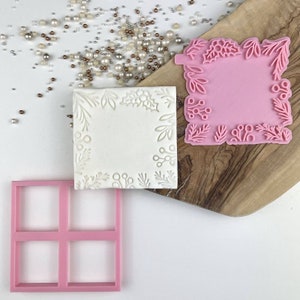 Peut inclure: Un tampon à biscuits carré rose avec une bordure florale, à côté d'un biscuit blanc avec un motif floral en relief. Un emporte-pièce carré rose et des perles décoratives sont également présents. Les articles sont sur une planche en bois et une surface blanche.