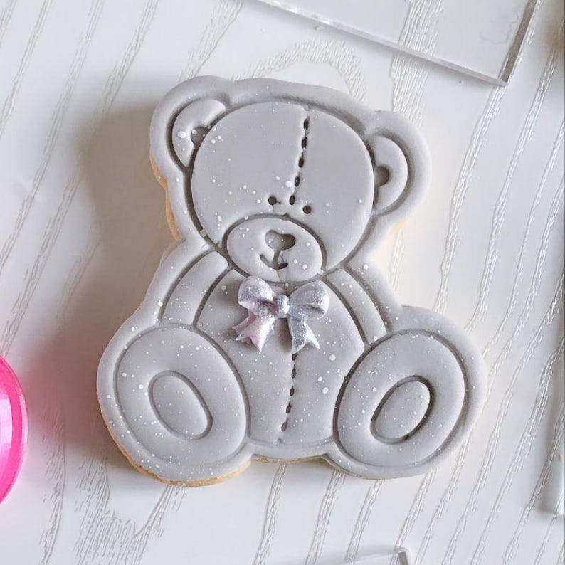 May include: A light gray teddy bear shaped cookie decorated with smooth icing. The bear features a stitched line down the center of its head and body, a small heart shaped nose, and a metallic silver bow tied around its neck with white speckles.