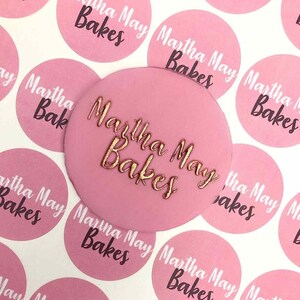 May include: Pink round cake topper with the text "Martha May Bakes" in gold lettering.