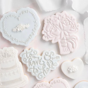 K&ouml;nnte beinhalten: Eine Auswahl an wei&szlig; glasierten Keksen mit aufwendigen Designs. Die Kekse umfassen Herzformen, einen Blumenstrau&szlig; und eine mehrst&ouml;ckige Torte. Einige Kekse zeigen den Text "To have & to hold" in eleganter Schrift. Die Kekse sind auf einer wei&szlig;en Oberfl&auml;che angeordnet.