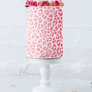以下が含まれることがあります： レオパード柄のデザインで飾られたピンクと白のケーキ。ケーキは白いケーキスタンドの上にあり、ピンクのバラの花びらで囲まれています。