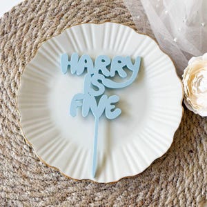 Può includere: Un cake topper azzurro con la scritta "HARRY IS FIVE" in un carattere giocoso. Il topper è posto su un piatto bianco con bordo dorato, suggerendo una festa di compleanno. Un fiore decorativo è visibile sullo sfondo.