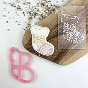 May include: A pink plastic cookie cutter shaped like a Christmas stocking. A white fondant Christmas stocking with a pink and yellow patterned design is on a wooden chopping board. A clear plastic stencil of a Christmas stocking is also on the chopping board.