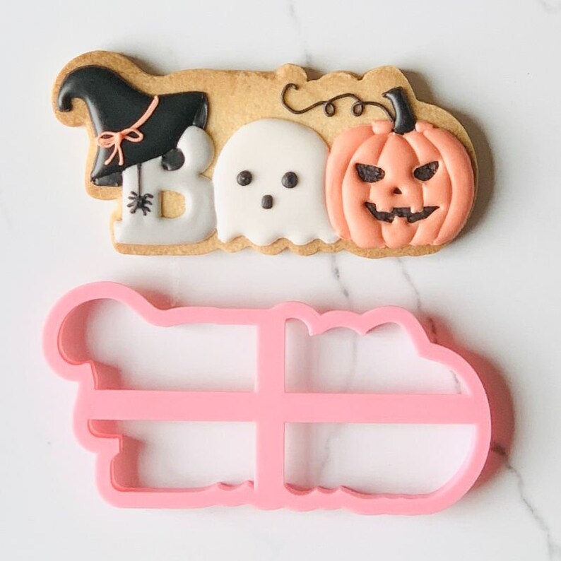 Puede incluir: Una galleta de color marr&oacute;n claro con la forma de la palabra BOO, decorada con un sombrero de bruja negro, un fantasma blanco y una calabaza color melocot&oacute;n, junto a un cortador de galletas de pl&aacute;stico rosa con el contorno del dise&ntilde;o.