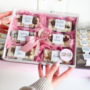 May include: A white gift box containing six rectangular cake slices wrapped in pink tissue paper. Each slice has a white label with text including Sticky Toffee, Walnut & Mocha, Cookies & Creme, Red Velvet, Carrot Cake, and Biscoff. A pink ribbon and a wooden spoon are tied on top, with a round Chell Bells Cakes logo sticker.