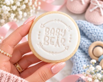 May include: A round sugar cookie with white fondant icing featuring the embossed text BABY BEAR inside a circular arrow design. The cookie is held against a background of baby shoes, a blue knitted blanket, and white flowers.