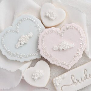 K&ouml;nnte beinhalten: Herzf&ouml;rmige Kekse mit wei&szlig;en Blumendesigns und hellblauem oder rosa Zuckerguss. Einige Kekse haben ein Blumengirlanden-Design am Rand. Ein rechteckiger Keks tr&auml;gt das Wort "bride" in silberner Schrift.