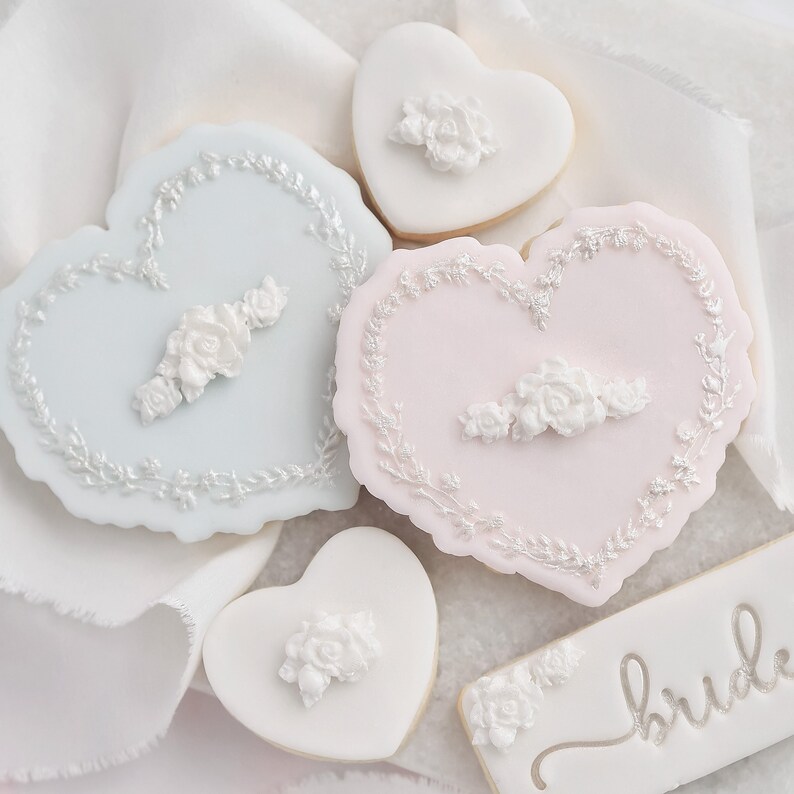 K&ouml;nnte beinhalten: Herzf&ouml;rmige Kekse mit wei&szlig;en Blumendesigns und hellblauem oder rosa Zuckerguss. Einige Kekse haben ein Blumengirlanden-Design am Rand. Ein rechteckiger Keks tr&auml;gt das Wort "bride" in silberner Schrift.